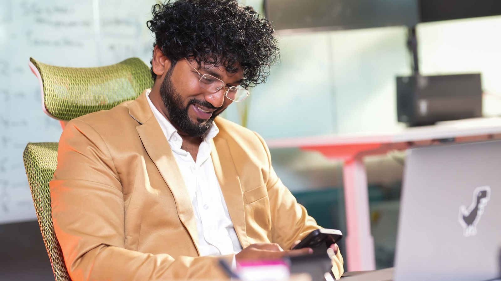 Smiling professional in a tan blazer typing a message on his smartphone at an office desk beside a laptop
